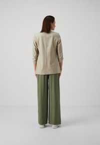 Blazer long beige clair à manches trois-quarts, associé à un pantalon vert olive ample et des baskets blanches. Design minimaliste et ajusté.