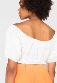 Top cropped branco de ombro a découvert com mangas bufantes, apresentando uma textura franzida e uma renda na bainha, combinado com calças laranja de cintura alta.