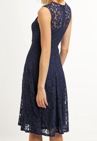 Navy blue lace dress with sleeveless design, fitted bodice, and flared skirt. Features a floral lace pattern and back zipper detail.