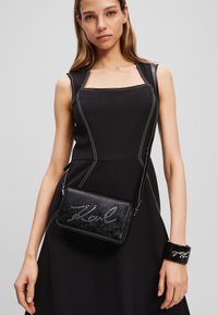 Čierna kožená crossbody taška so študovaným nápisom, elegantným dizajnom a striebornými akcentmi, skombinovaná s priliehavými čiernymi šatami a zodpovedajúcim náramkom.