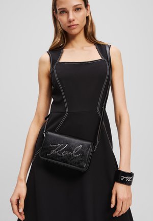 Čierna kožená crossbody taška so študovaným nápisom, elegantným dizajnom a striebornými akcentmi, skombinovaná s priliehavými čiernymi šatami a zodpovedajúcim náramkom.