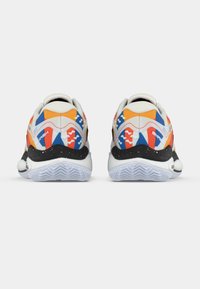 Krāsainu sporta apavu pāris ar oranžām, zilām, sarkanām un baltām dizaina schemām, melnām raibām apakšām un caurspīdīgām viļņotām zolem, skatīts no aizmugures.