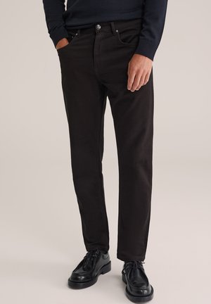 Homme portant un pantalon marron foncé et des chaussures en cuir noir, avec une main dans la poche et l'autre reposant à ses côtés.