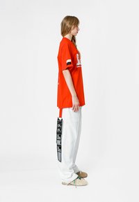 Camiseta roja de gran tamaño con estampado gráfico, combinada con pantalones holgados blancos que tienen una franja negra y un detalle de etiqueta. Zapatos marrón completan el conjunto.