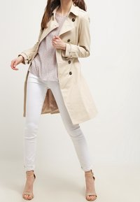 Trench beige con botones de doble botonadura, mangas largas y un corte ligeramente oversize, combinado con jeans skinny blancos y tacones beige.