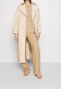 Beige läder trenchcoat med stora slag och guldknappar, kombinerad med en ljus beige topp och vida byxor, bärs med bruna slidesandaler.