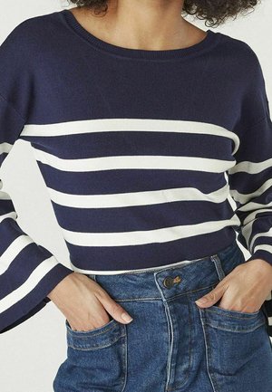 Frau trägt einen dunkelblauen Pullover mit weißen horizontalen Streifen und weiten Ärmeln, Hände in den Vorderentaschen einer hoch taillierten blauen Jeans.