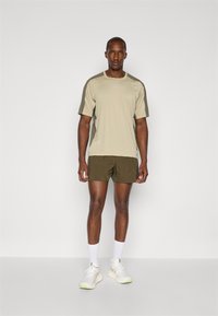 Beige athletic t-shirt with olive green accents, paired with dark green shorts. Model wears white sneakers and ankle-length white socks.