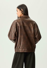 Femme aux longs cheveux noirs portant de grandes boucles d'oreilles en or et une veste en cuir marron ample, tournée de côté contre un fond uni clair.