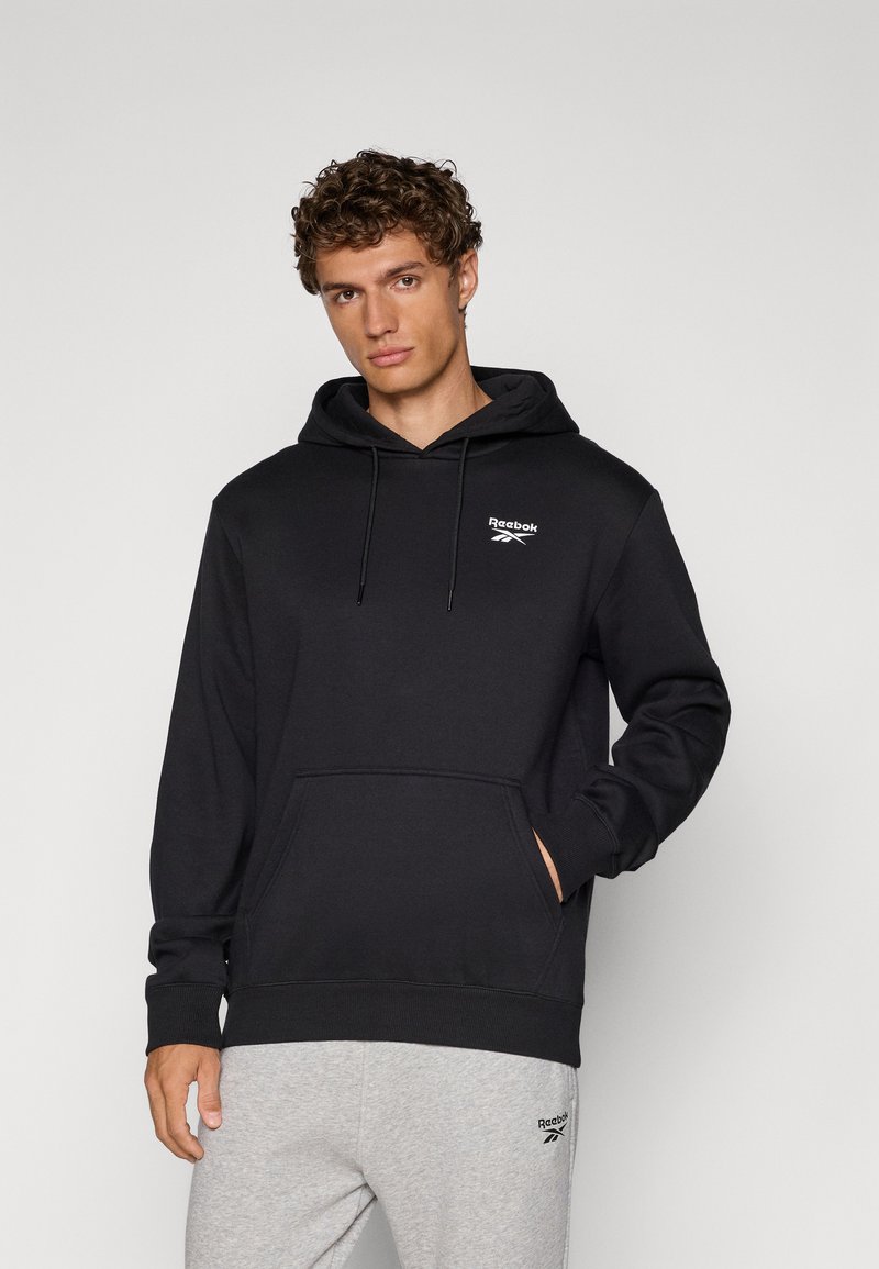 Čierna mikina s kapucňou a predným vreckom, so šnúrkami na kapucni a logom Reebok na hrudi. Je kombinovaná so sivými teplákmi, ktoré majú logo na stehne.