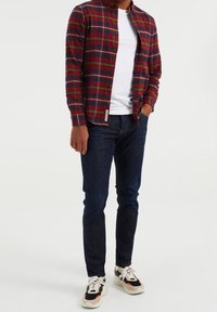 Mies, który ma na sobie czerwoną i niebieską koszulę w kratę na białym T-shircie, ciemnoniebieskie dżinsy oraz białe sportowe buty z czarnymi i czerwonymi detalami, stoi z jedną ręką w kieszeni.