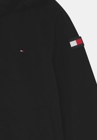 Czarna kurtka z małym prostokątnym logo w czerwonym, białym i granatowym kolorze na klatce piersiowej oraz naszywką na górnej części rękawa, prezentowana na białym tle.