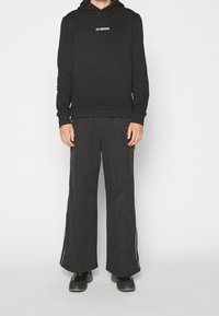 Felpa nera con cappuccio, tasca frontale e logo bianco; abbinata a pantaloni neri a gamba larga con cerniere laterali e tessuto liscio.