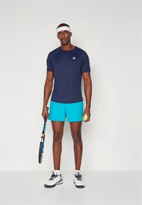 T-shirt atlético azul-marinho com um logótipo, calções azul brilhante, segurando uma raquete e uma bola de ténis, usando uma faixa branca na cabeça e sapatos desportivos.