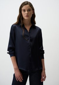 Navyblauwe zijden shirt met lange mouwen, knoopsluiting aan de voorkant en een gebogen zoom; gladde textuur en subtiele glans. Draagt het met donkere jeans.