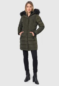 Olivgrüner Puffercoat mit einer schwarzen Kunstfellkapuze, vorderen Knöpfen, seitlichen Taschen und gestepptem Design; kombiniert mit schwarzen Hosen und hohen Stiefeln.