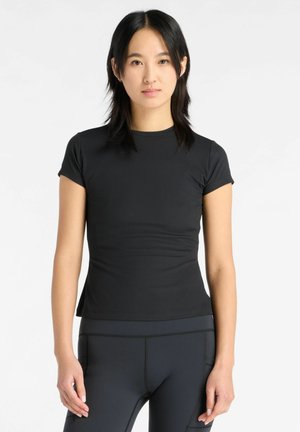 Ung kvinde med sort, glat hår iført en tætsiddende sort ribbet kortærmet top og mørke leggings, står mod en hvid baggrund.