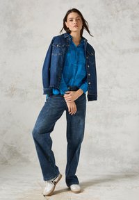 Chaqueta de mezclilla con dos bolsillos en el pecho, vestida sobre una camisa azul con estampado. Jeans sueltos de color azul oscuro y zapatillas blancas completan el conjunto.