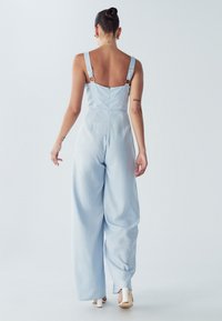 Jasnoniebieski kombinezon z szerokimi nogawkami, z regulowanymi ramiączkami i zamkiem błyskawicznym z tyłu. Miękka tkanina o gładkiej teksturze i minimalistycznym wzornictwie.