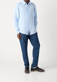 Camicia di lino azzurra chiara con colletto button-down e taschino sul petto, abbinata a jeans blu scuro e sneakers nere. Adatta a un look casual.