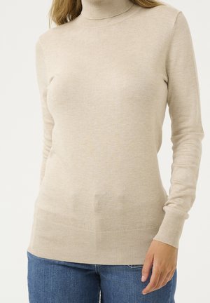 Femme portant un pull à col roulé beige à manches longues associé à un jean bleu, debout devant un fond clair uni.