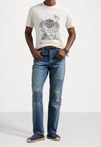 Kremowa koszulka graficzna z motywem palm, zestawiona z jasnoniebieskimi spodniami dżinsowymi z przetarciami. Niebieskie espadryle z beżowymi akcentami dopełniają stylizację.