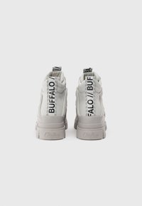 Korkean varren tennarit valkoisesta synteettisestä materiaalista, joissa on pehmustetut varsitukat, paksut beiget pohjat ja mustat logot yksityiskohtina kannassa ja takanauhassa.