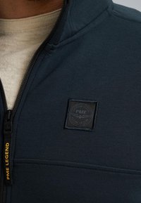 Donkerblauwe ritsjack met een gestructureerde stof, voorzien van een vierkante zwarte logopatch en een gele trekband met het label "PME LEGEND."