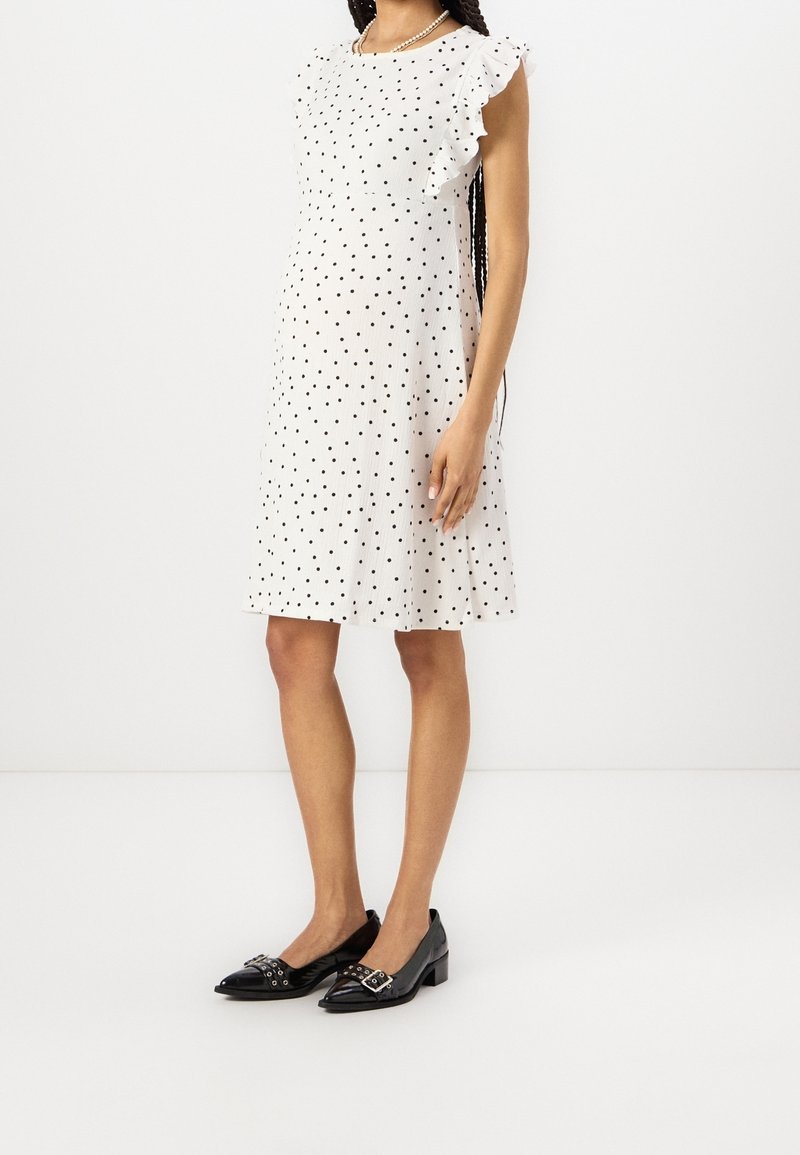 Valkoinen pilkullinen mekko, jossa on röyhelöiset hartiaosat ja rento istuvuus, yhdistetty mustiin, solkikoristeellisiin teräväkärkisiin tasapohjaisiin kenkiin.