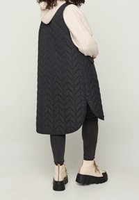Gilet long matelassé noir avec un motif en chevron, fentes latérales, porté sur un sweat à capuche léger, associé à un jean skinny noir et des bottines beige.