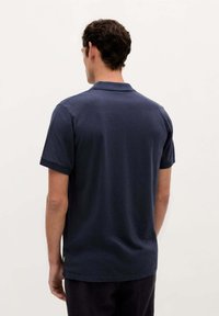 Marineblaue Polohemd mit kurzen Ärmeln, ausgestattet mit einem klassischen Kragen und einem subtilen strukturierten Stoff. Der Saum hat ein kleines Etikett an der Seite.