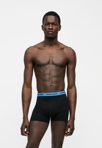 Άνδρας χωρίς πουκάμισο με μυώδες σώμα, φορώντας μαύρα ανδρικά εσώρουχα με μπλε μέση Björn Borg, στέκεται μπροστά σε απλό ανοιχτό φόντο.