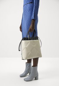 Femme portant une robe côtelée bleue et des bottines bleu clair tenant un grand sac matelassé crème et noir avec le logo CK.