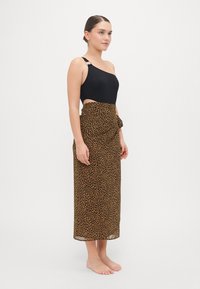 Maillot de bain noir à une épaule avec un détail en anneau, associé à une jupe portefeuille à imprimé léopard marron avec un nœud à la taille.