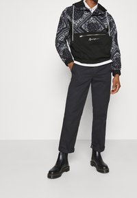 Man i svart mönstrad tröja med ficka framtill, mörka uppvikta byxor och svarta läderstövlar som står mot en enkel vit bakgrund.