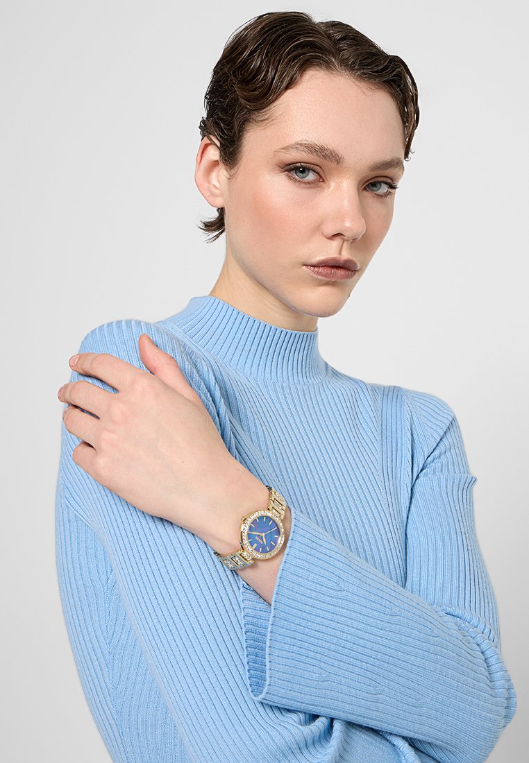 Montre dorée avec un cadran bleu, face ronde et bracelet métallique texturé, portée sur un poignet au-dessus d'un pull à côtes bleu clair.