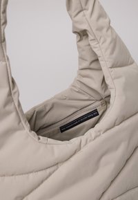 Borsa puffa beige con una morbida texture trapuntata. Il materiale principale è composto da reti da pesca riciclate. Etichetta interna visibile, che indica la fonte del materiale.