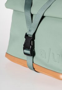 Zaino in materiale morbido verde con base in pelle marrone chiaro, dotato di una cintura nera con fibbia e dettaglio del logo impresso sul lato.