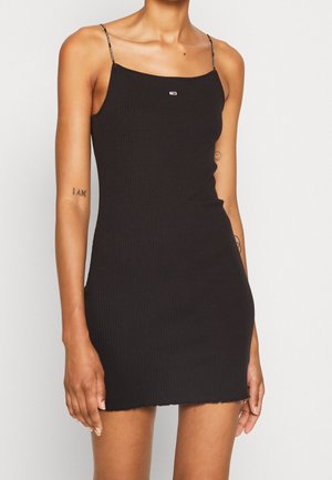 Femme portant une mini-robe noire côtelée ajustée avec fines bretelles spaghetti et petit logo rectangulaire sur la poitrine.