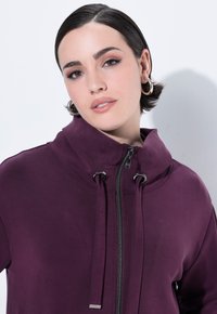 Chaqueta morada con cremallera, que presenta un cuello alto, cordones ajustables y una textura suave y elástica, con herrajes metálicos en la cremallera.