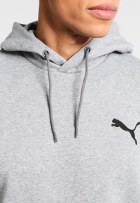 Grå hoodie av mjukt material med känguruficka, dragsko på huvan och en svart Puma-logotyp på bröstet.