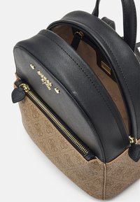 Kompakti kaksivärinen Guess-reppu, jossa on teksturoitu musta yläosa, beige monogrammifarkun pohja, kultaiset vetoketjut ja logovarusteet, osittain auki, paljastaen sisäpuolen.
