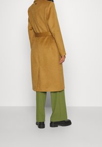 Lång, figursydd kappa i senapsgul ullblandning, med skärp i midjan, med en ren design och slits bak. Kombineras med gröna vida byxor.