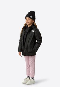 Schwarze wasserdichte Jacke mit Kapuze, Reißverschluss und zwei Fronttaschen. Kombiniert mit einer schwarzen Mütze, pinken Hosen und weißen Sneakers.