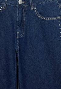 Temno modre kavbojke iz jeansa s srebrnimi zakovicami ob sprednjih žepih, kontrastnim šivom in klasično zasnovo s petimi žepi.