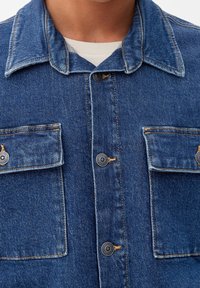 Chaqueta de mezclilla azul con cierre de botones, dos bolsillos en el pecho con solapa y un cuello clásico. Acentos de costura visibles en la tela.