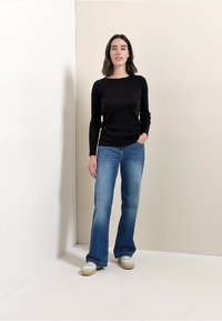 Mustassa ribattussa pitkähihaisessa paidassa, jota on täydennetty vaaleansinisillä leveälahkeisilla farkuilla. Valkoiset tennarit, joissa on beige pohja. Yksinkertainen tausta neutraaleissa väreissä.