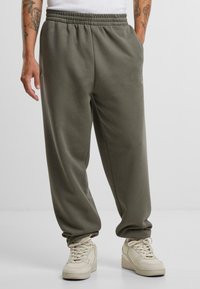 Lightweight, olive green sweatpants with an elastic waistband, relaxed fit, side pockets, and subtle logo accent. Paired with beige sneakers.