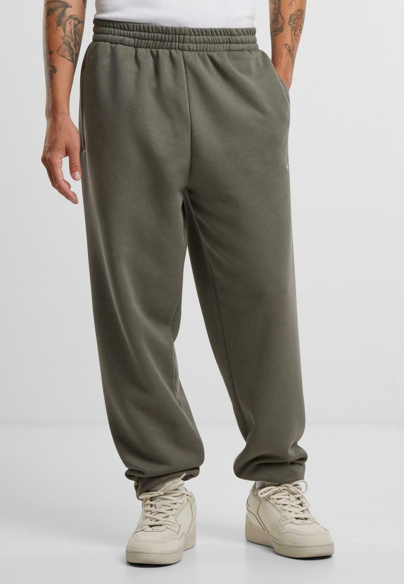 Lightweight, olive green sweatpants with an elastic waistband, relaxed fit, side pockets, and subtle logo accent. Paired with beige sneakers.