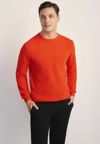 Czerwony sweter o fakturze z okrągłym dekoltem i długimi rękawami, w połączeniu z czarnymi spodniami. Projekt zawiera prążkowane mankiety i dół.
